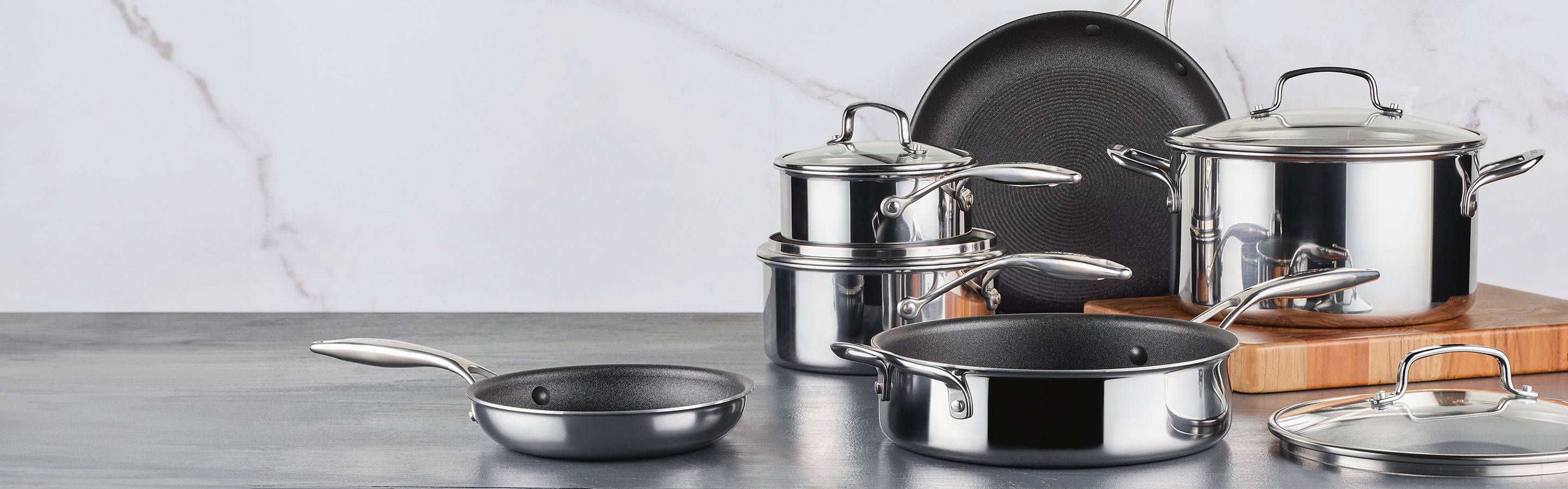A set of stainless steel cookware, including saucepans, frying pans, and pots with lids, is arranged on a grey countertop with a marble background.