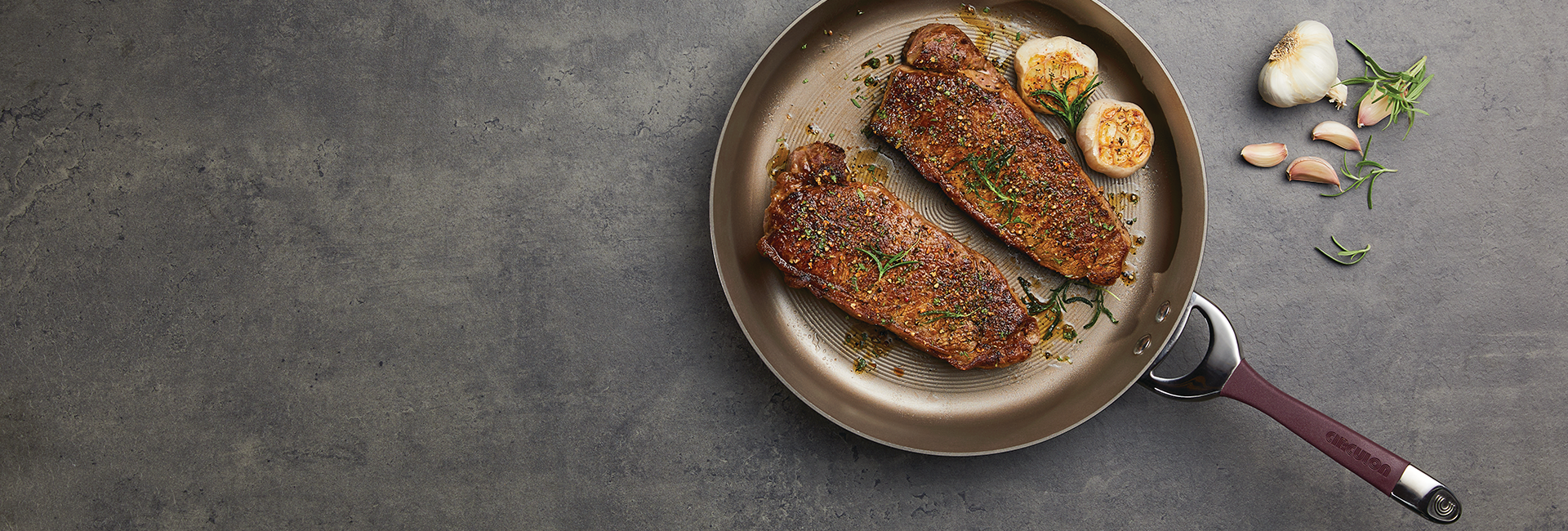 Two cooked steaks with herbs and garlic cloves in a frying pan on a gray surface, with additional garlic and rosemary beside the pan.