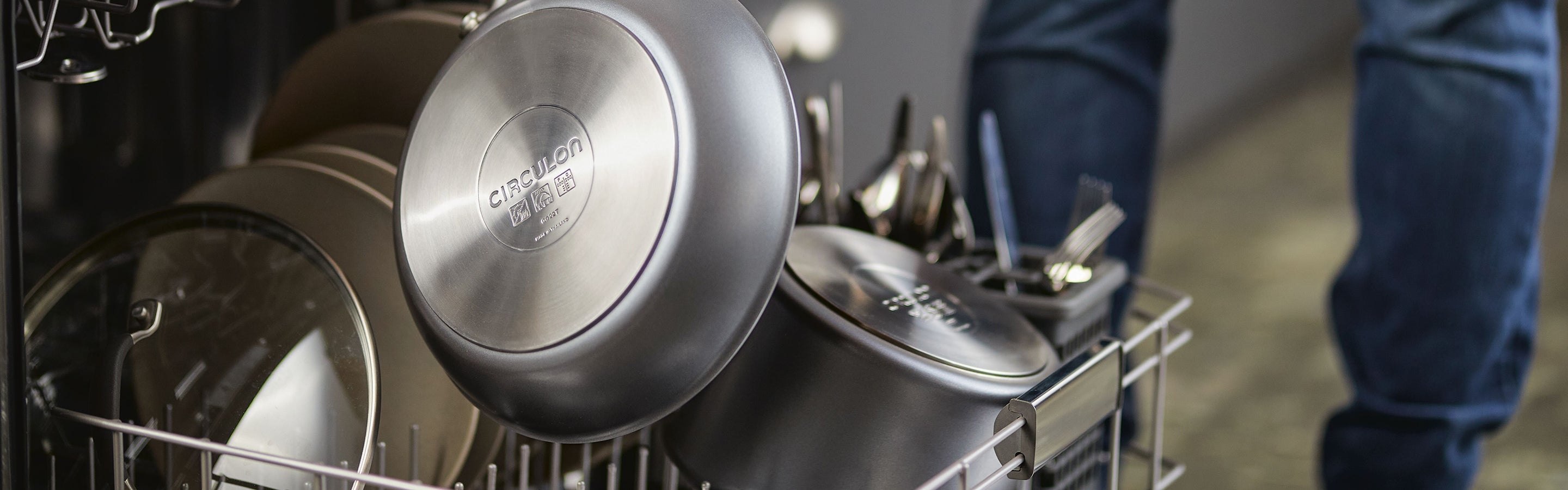 Open dishwasher with stainless steel pots and pans loaded on the bottom rack, along with utensils and a glass lid.