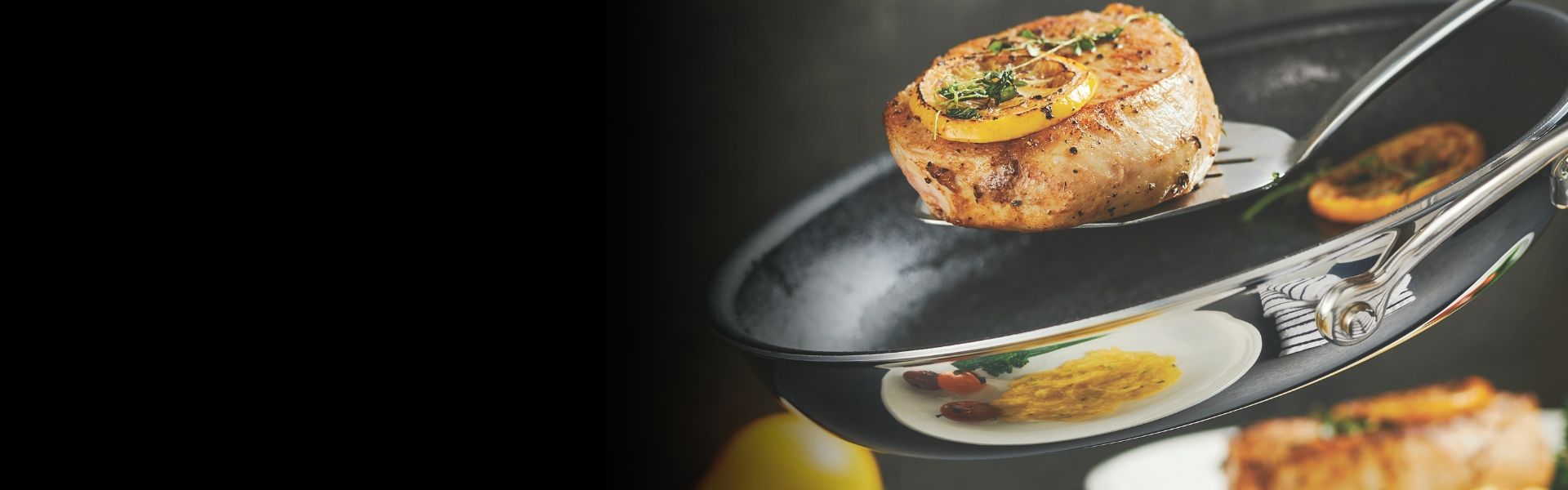 A seasoned piece of meat on a spatula is being lifted from a pan, with a slice of lemon and herbs garnishing it. A plate with sides is visible in the background.