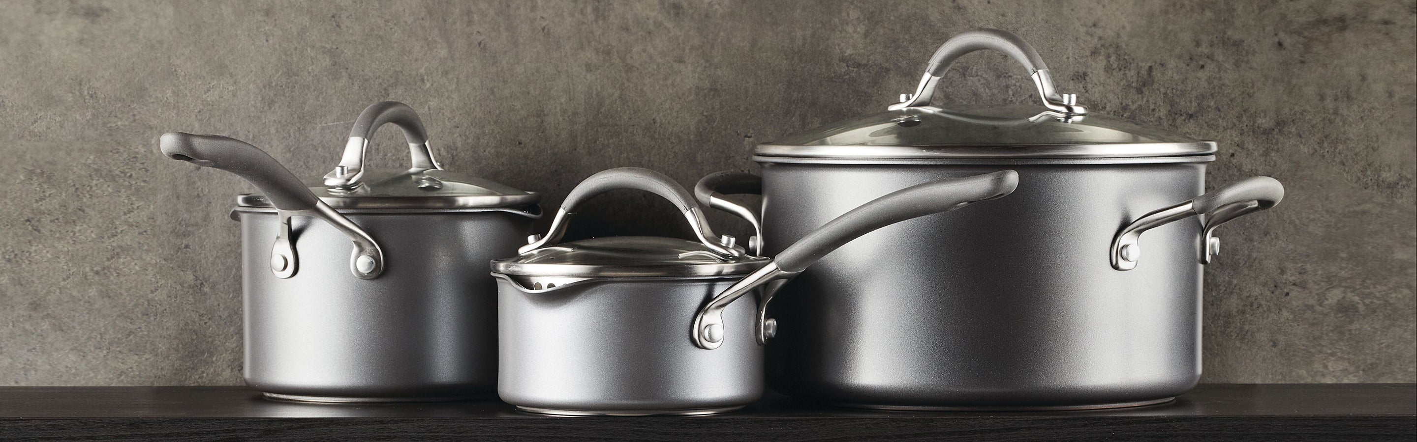Three silver metal pots with lids and handles are displayed in a row against a gray textured background.