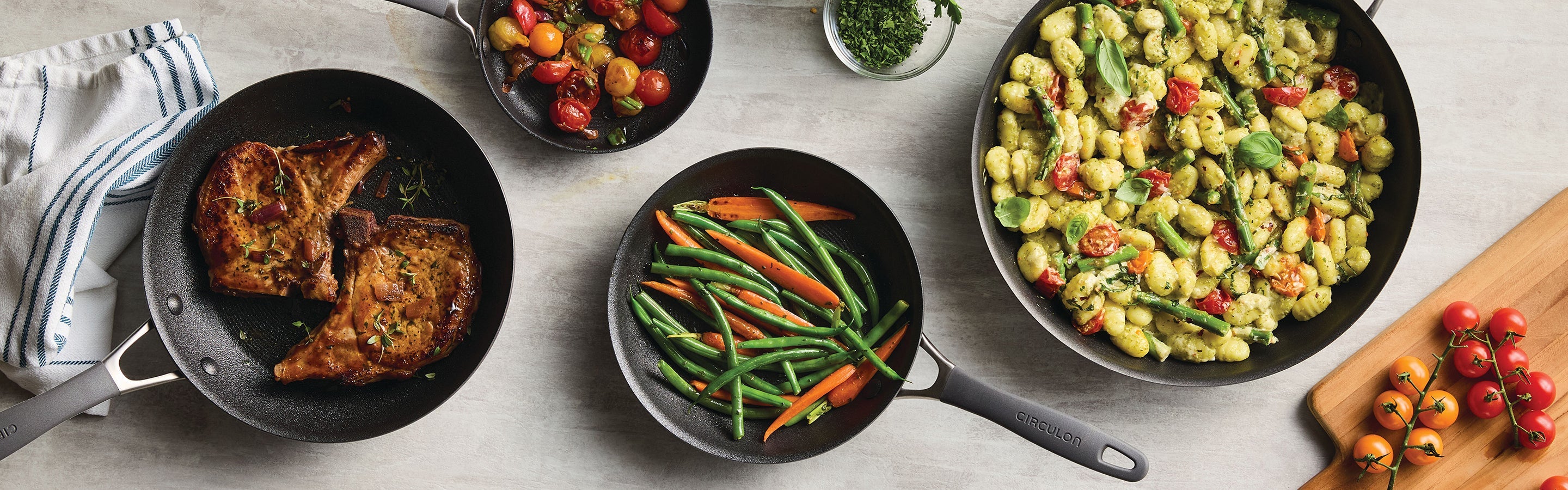 Two seared pork chops with herbs are being cooked in a frying pan; a person is using tongs to turn one chop.