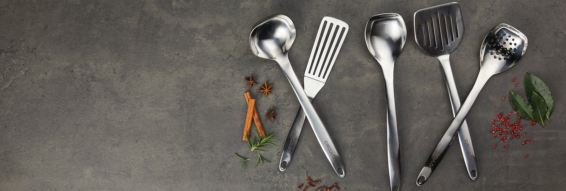 Five stainless steel kitchen utensils lie on a gray surface surrounded by herbs and spices.