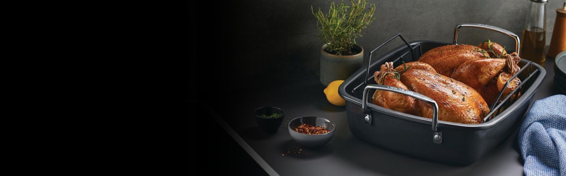 Two roasted chickens in a black roasting pan with herbs, spices, and a lemon nearby on a dark kitchen counter.