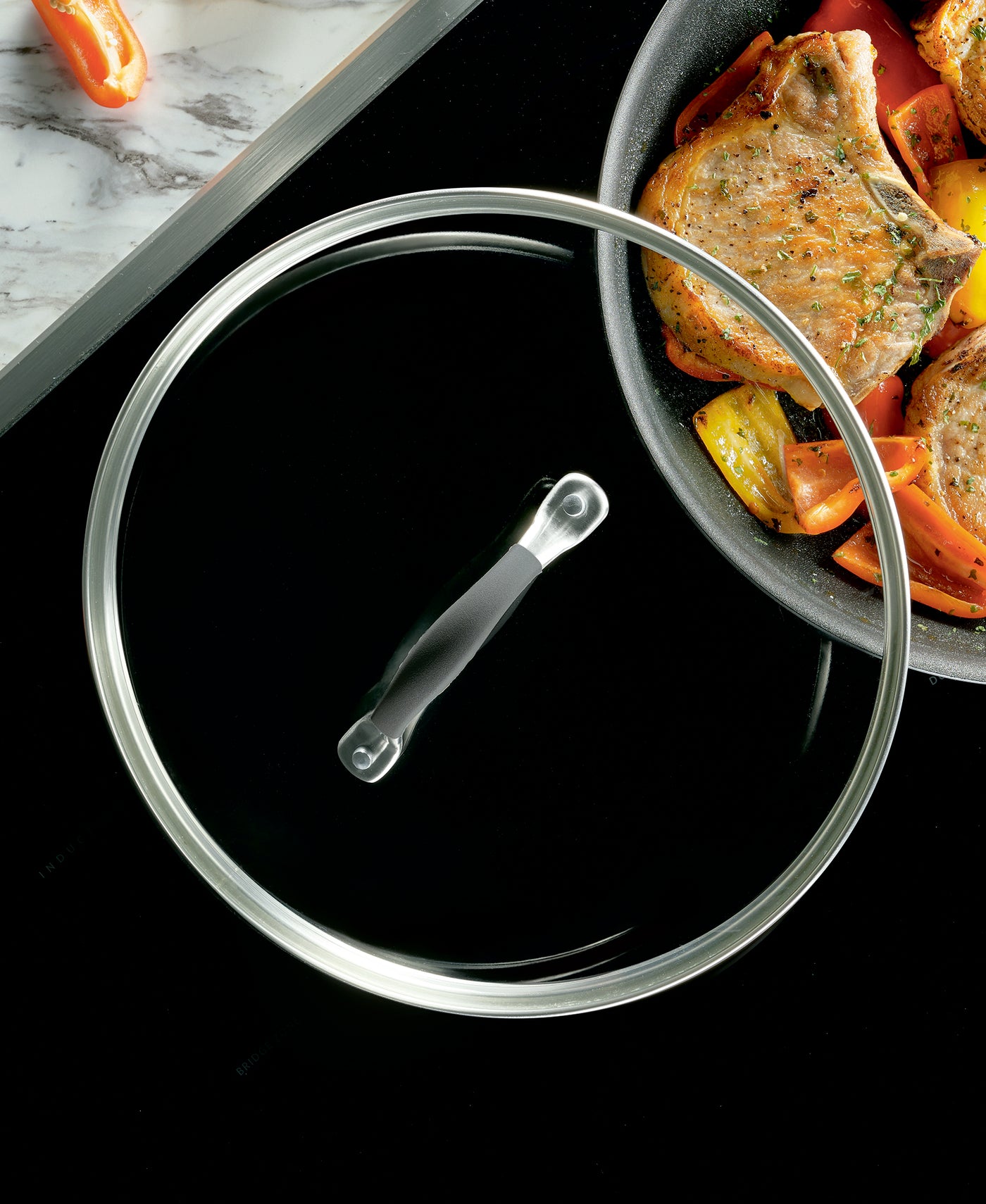 Glass lid next to a pan with cooked meat and vegetables.