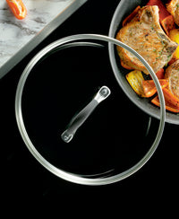 Glass lid next to a pan with cooked meat and vegetables.