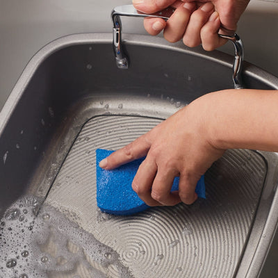 A hand scrubs a baking tray with a blue sponge in soapy water.