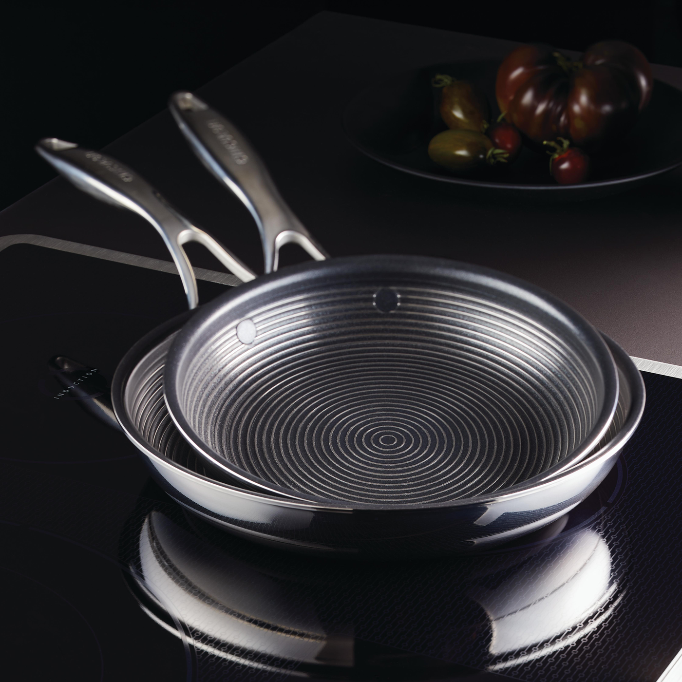 Two stacked frying pans on a stovetop with tomatoes in the background.