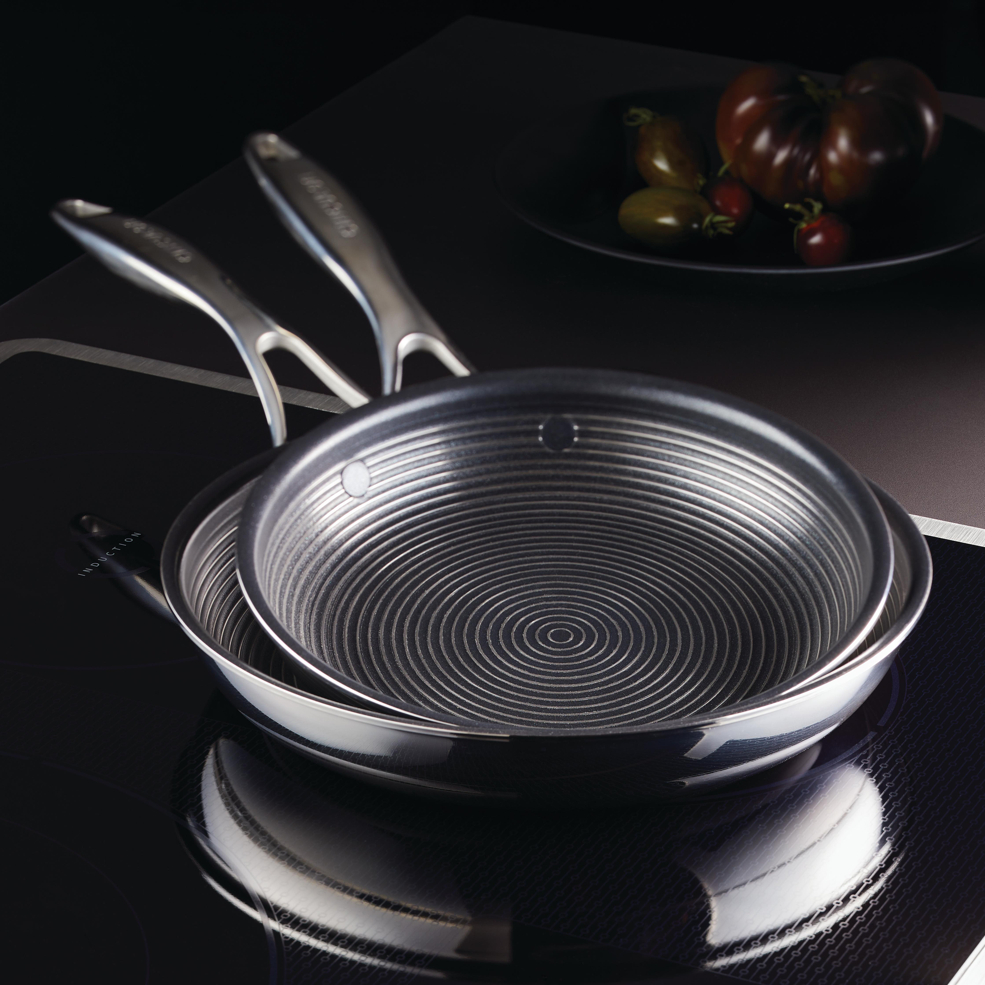 Two stacked frying pans on a stovetop with tomatoes in the background.