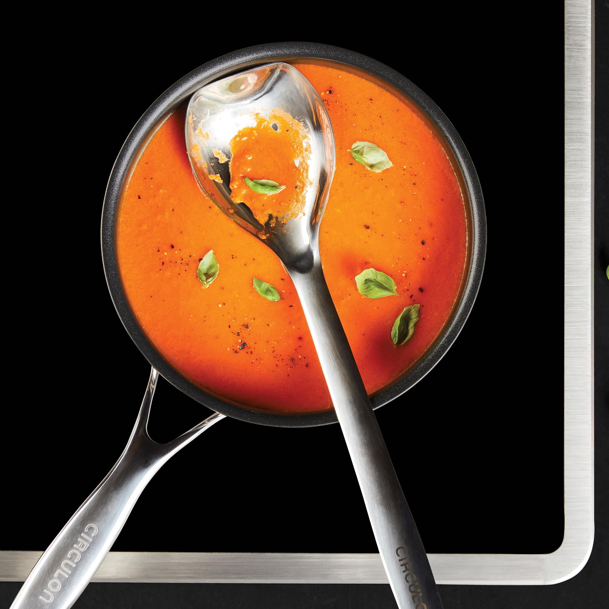 Tomato soup with basil leaves in a pot, with a ladle on a black background.