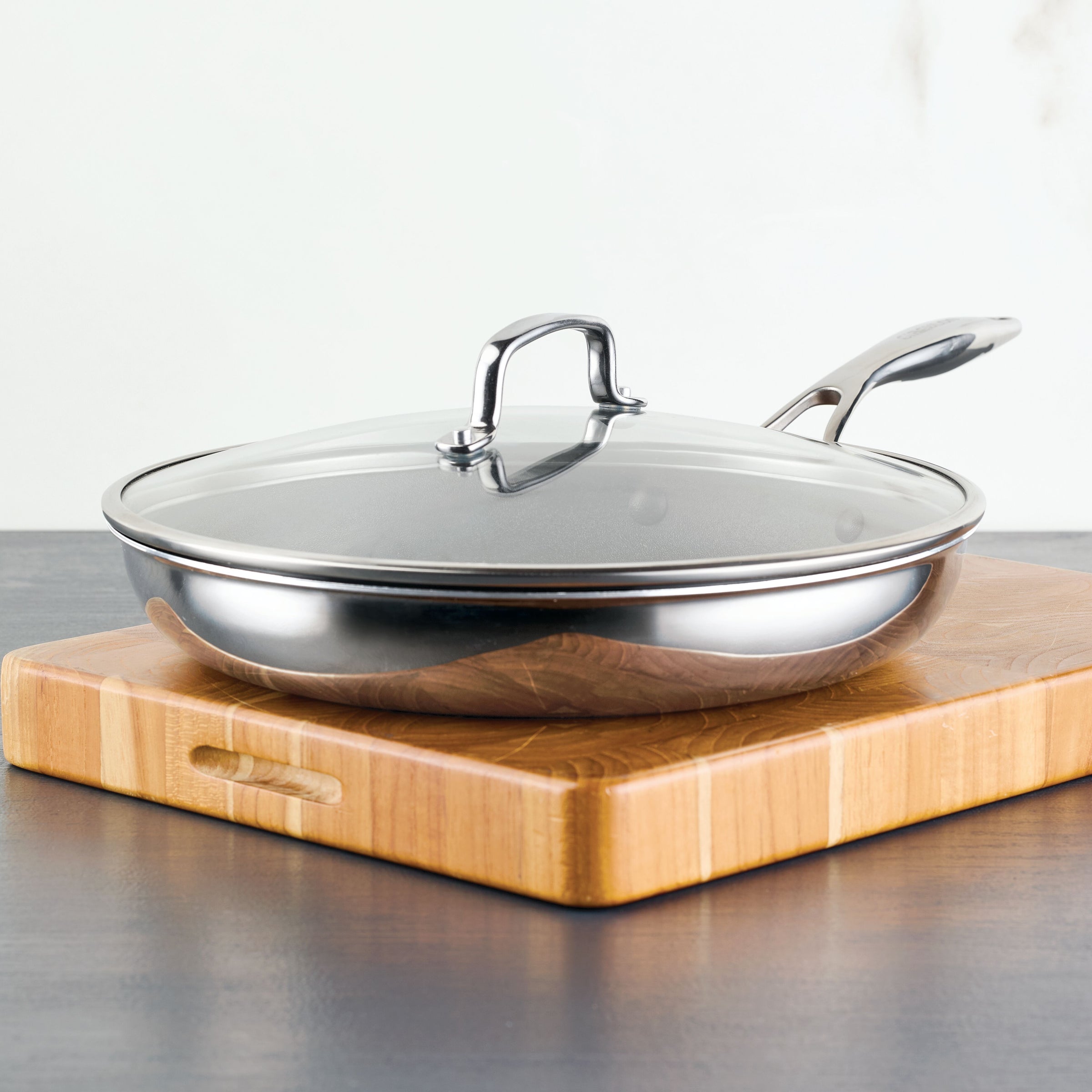 A stainless steel frying pan with a glass lid on a wooden cutting board.
