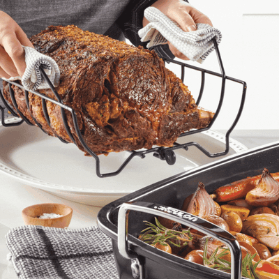 Person lifting a cooked roast from a pan, with vegetables in another tray.