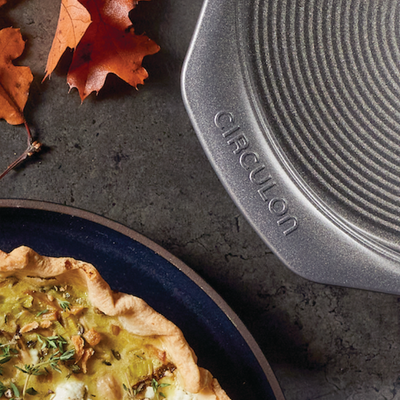 A quiche next to a circular baking tray and autumn leaves on a dark surface.