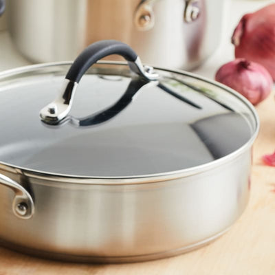 Stainless steel pot with a glass lid on a wooden surface, onions nearby.