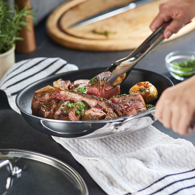 Person using tongs to cook steak in a pan with herbs and potatoes.