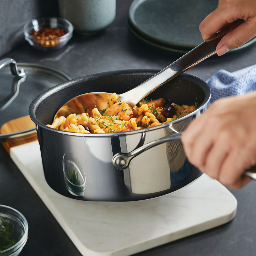 A person stirs pasta and vegetables in a pot on a kitchen counter.
