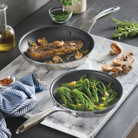 Two frying pans with meat and broccoli on a marble countertop, herbs nearby.