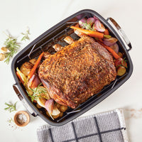 Juicy roasted meat with vegetables rests in the Roasting Pan: Nonstick Roaster Pan with Easy Serve Rack, elegantly presented on a white surface.