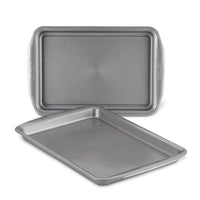 Displayed on a white background, the 2-Piece Nonstick Cookie Sheet Set features two metal baking trays, with one slightly tilted. These oven-safe pans guarantee effortless baking and easy cleanup.